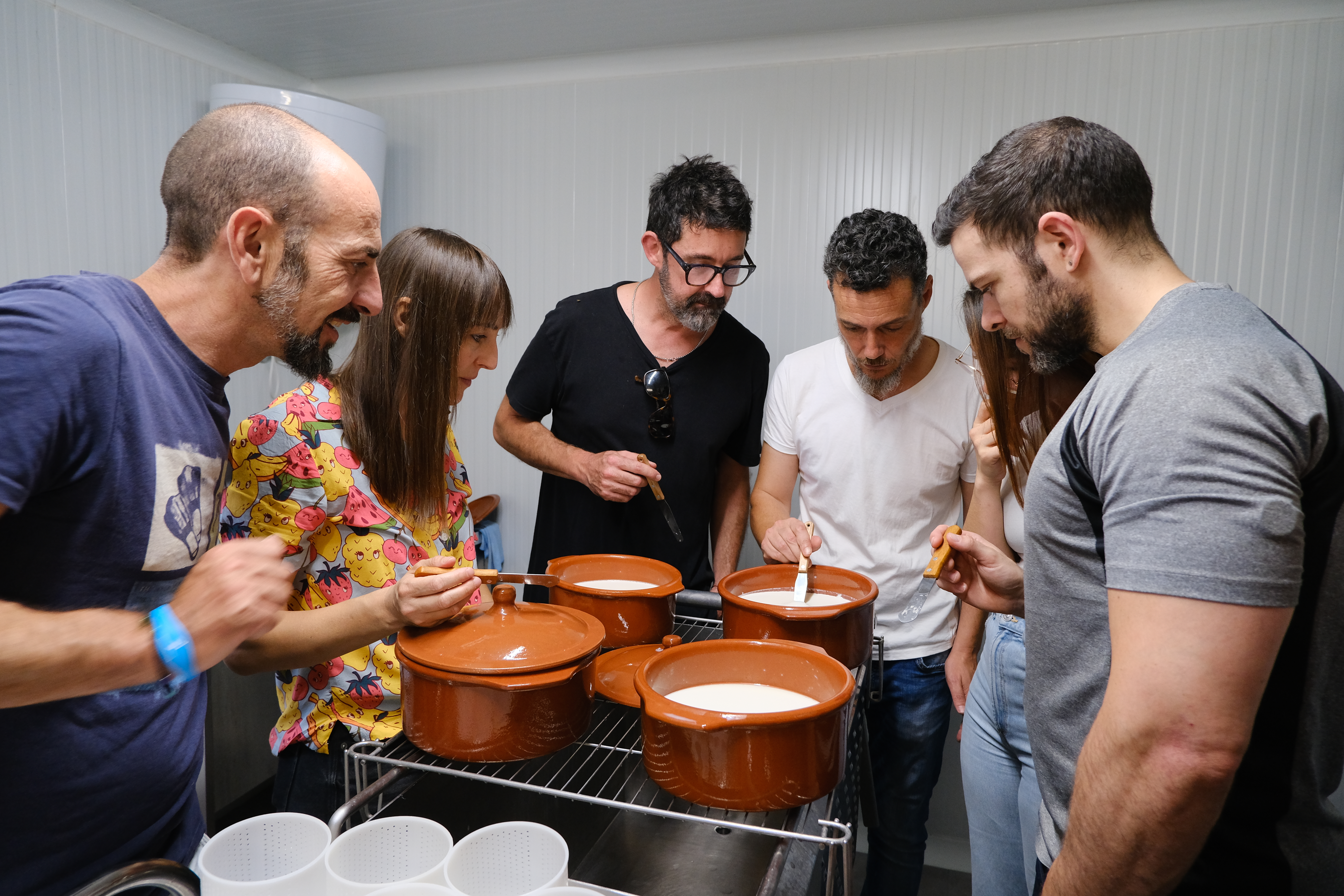 Un grupo de personas reunidas alrededor de ollas de cerámica durante un taller de elaboración de queso, observando y removiendo la leche con curiosidad y atención.